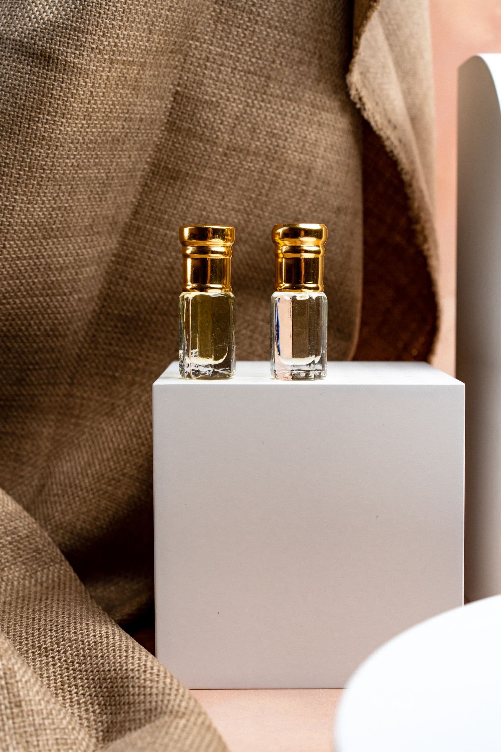 Ambroisie Dorée — two small glass perfume oil bottles with gold caps on a white plinth, set against a warm linen backdrop
