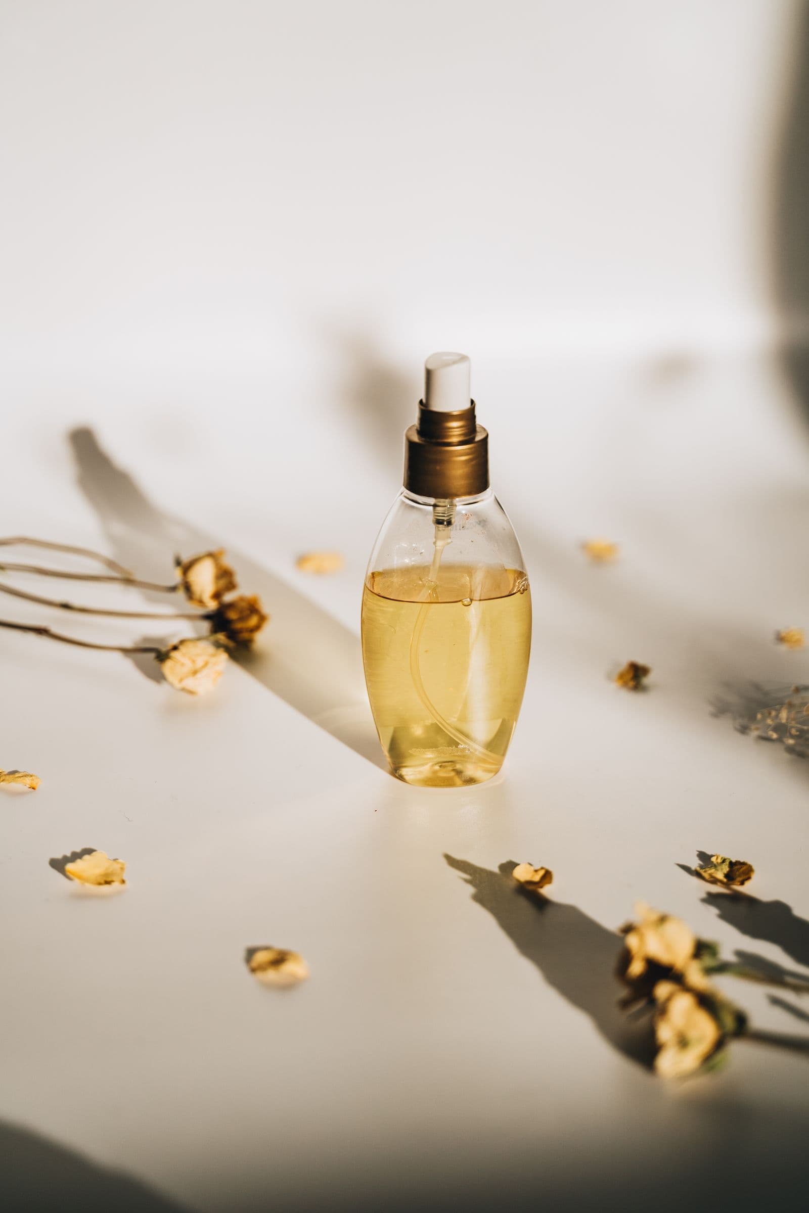 Pearl Bloom — a teardrop-shaped glass perfume bottle with a gold-and-white cap, golden liquid inside, surrounded by scattered dried petals