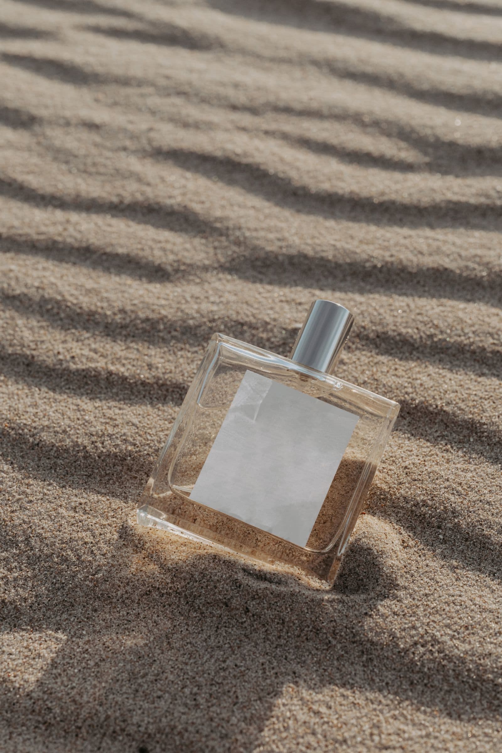 Safran Céleste — a square glass perfume bottle with a silver cap and blank white label, standing upright in wind-rippled sand dunes